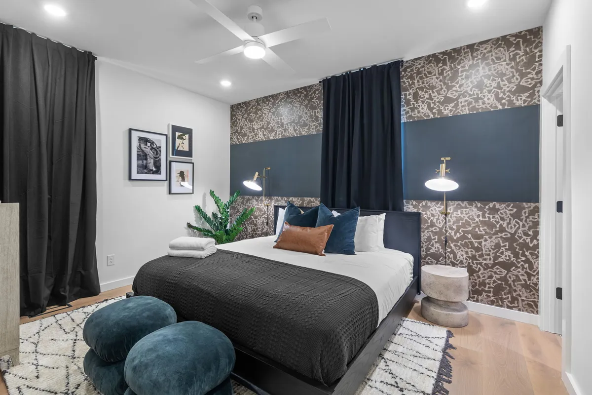 A stylish bedroom with a neatly made bed, decorative pillows, artful wall decor, plants, and soft lighting. The design is both cozy and modern, offering a warm environment for restful sleep or leisurely mornings.