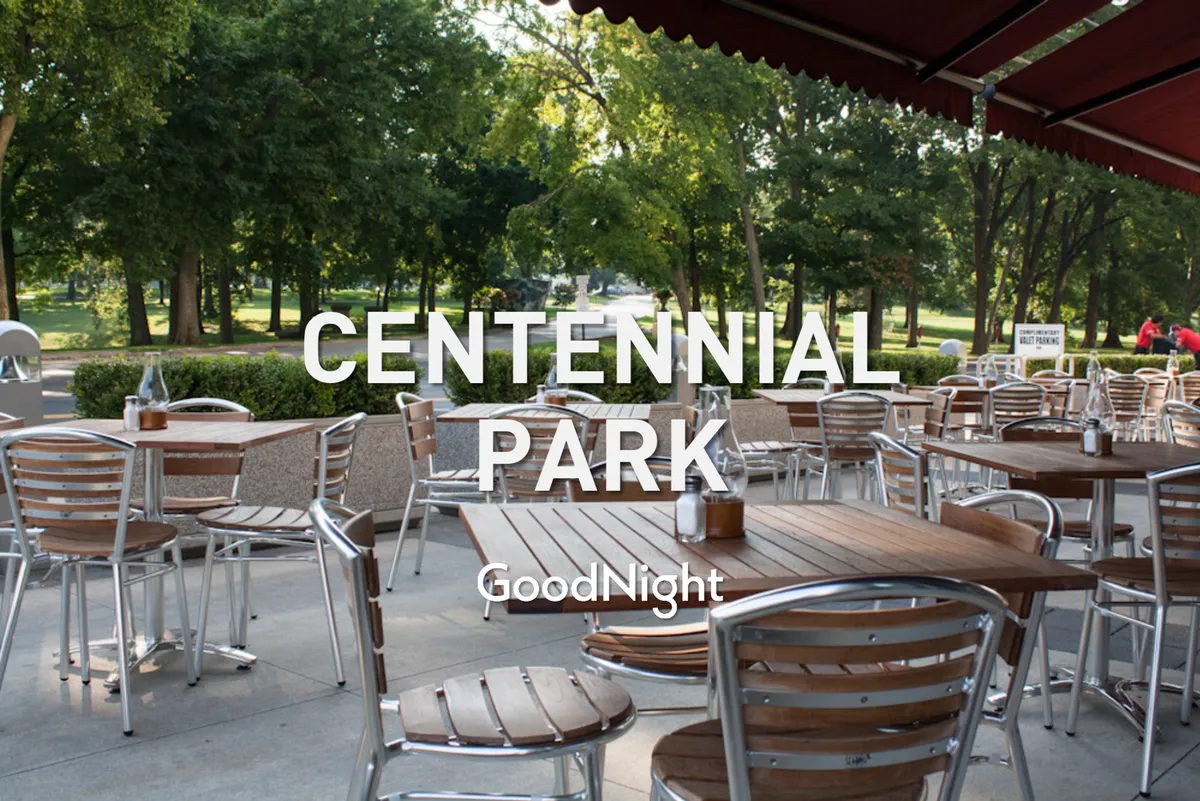 An inviting outdoor dining area featuring wooden tables, metal chairs, and landscaped greenery, with a distant park scene that hints at relaxation and social gatherings in a natural setting.