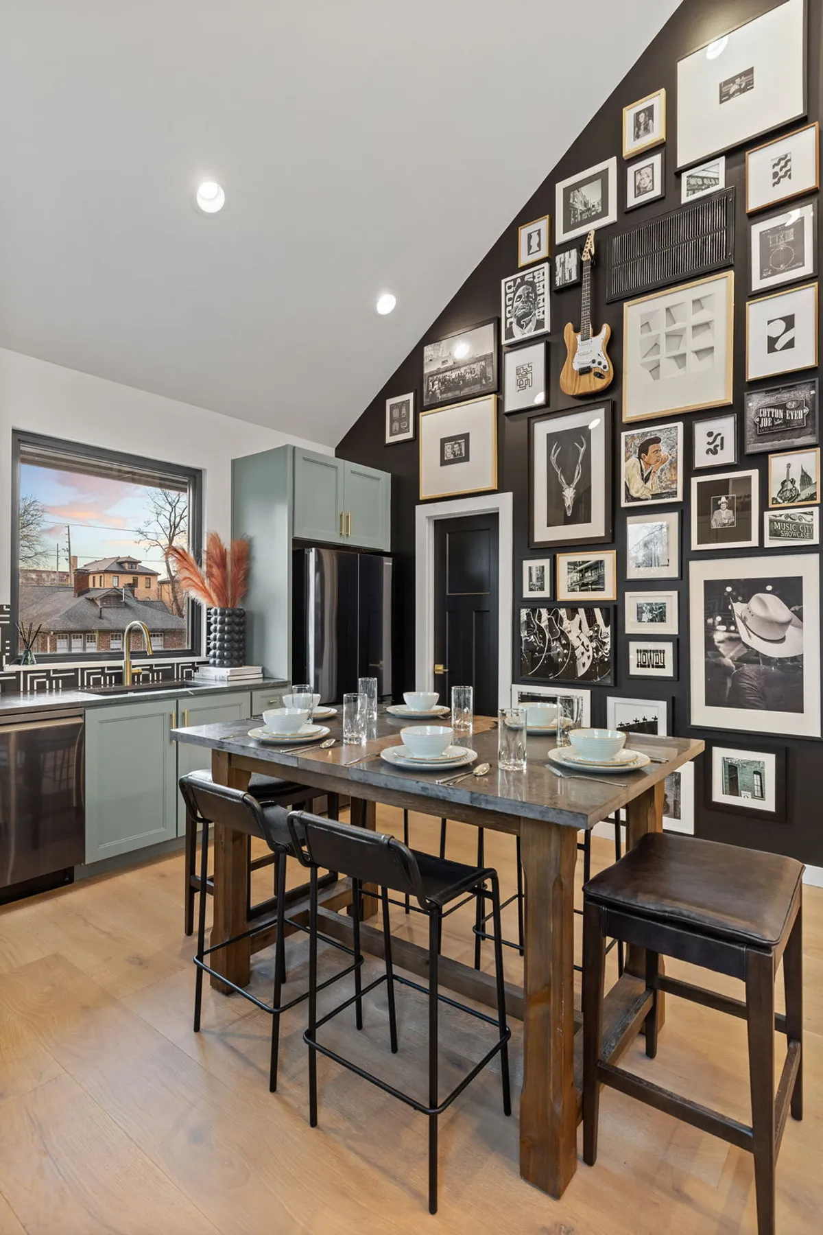 This space highlights a kitchen island with seating, a gallery wall of framed local art and photos, and a large window offering a glimpse outside. The overall atmosphere is creative, relaxed, and conducive to both cooking and socializing.