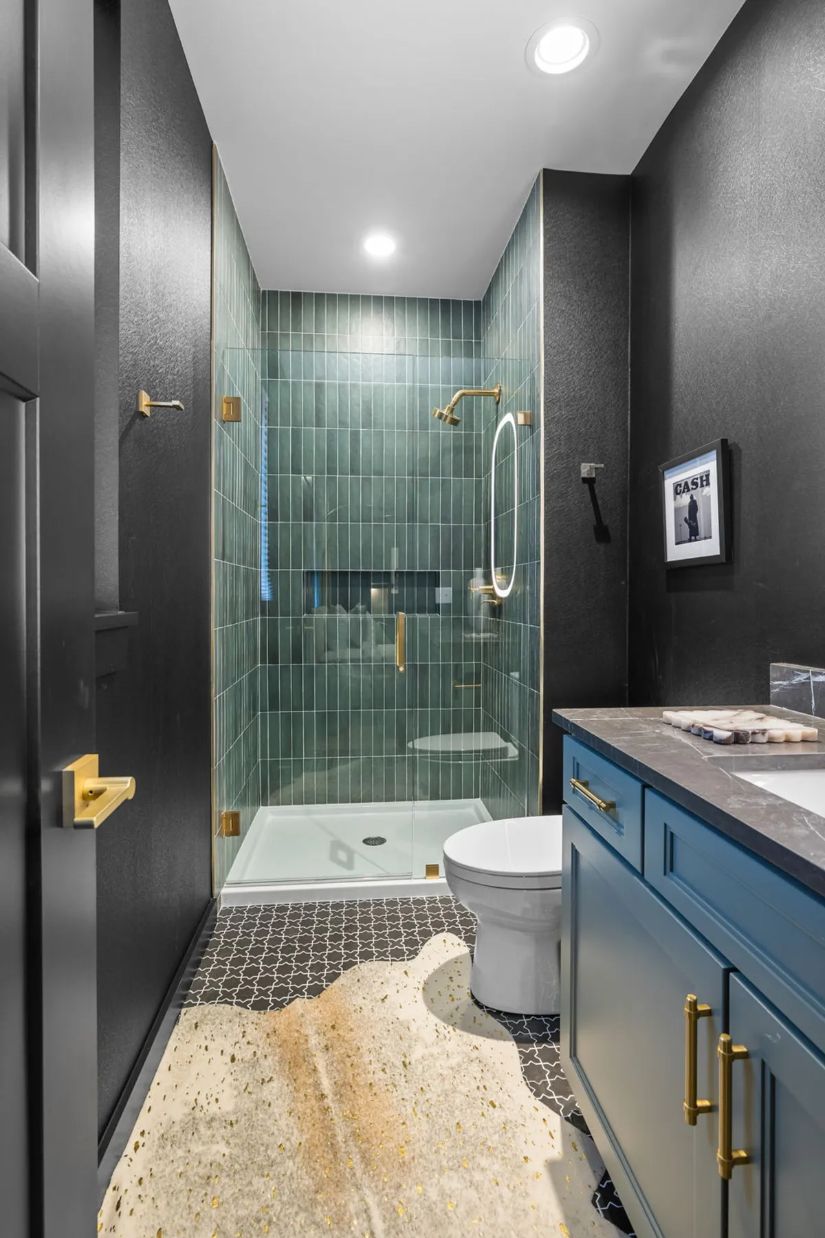 A sophisticated en-suite bathroom featuring a green-tiled shower, gold fixtures, a blue vanity with a black accent wall, and a decorative rug. The space exudes serenity and luxury, perfect for a private self-care retreat.