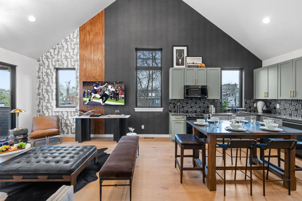This image presents a spacious, open-plan living and dining area illuminated by large windows. The stylish sectional sofa faces a large flat-screen TV, while a modern dining table is set for a meal. Unique textured walls and abundant natural light give the space a welcoming, contemporary vibe.