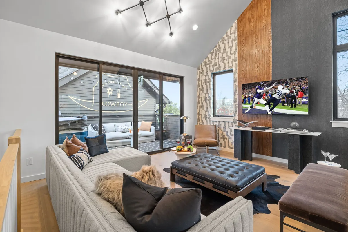 A spacious, sunlit living room featuring a sleek modern sofa set against a decorative wall, large windows letting in natural light, a prominent television, and a stylish coffee table. The space feels inviting, perfect for socializing, relaxing, or watching the big game with friends and family.