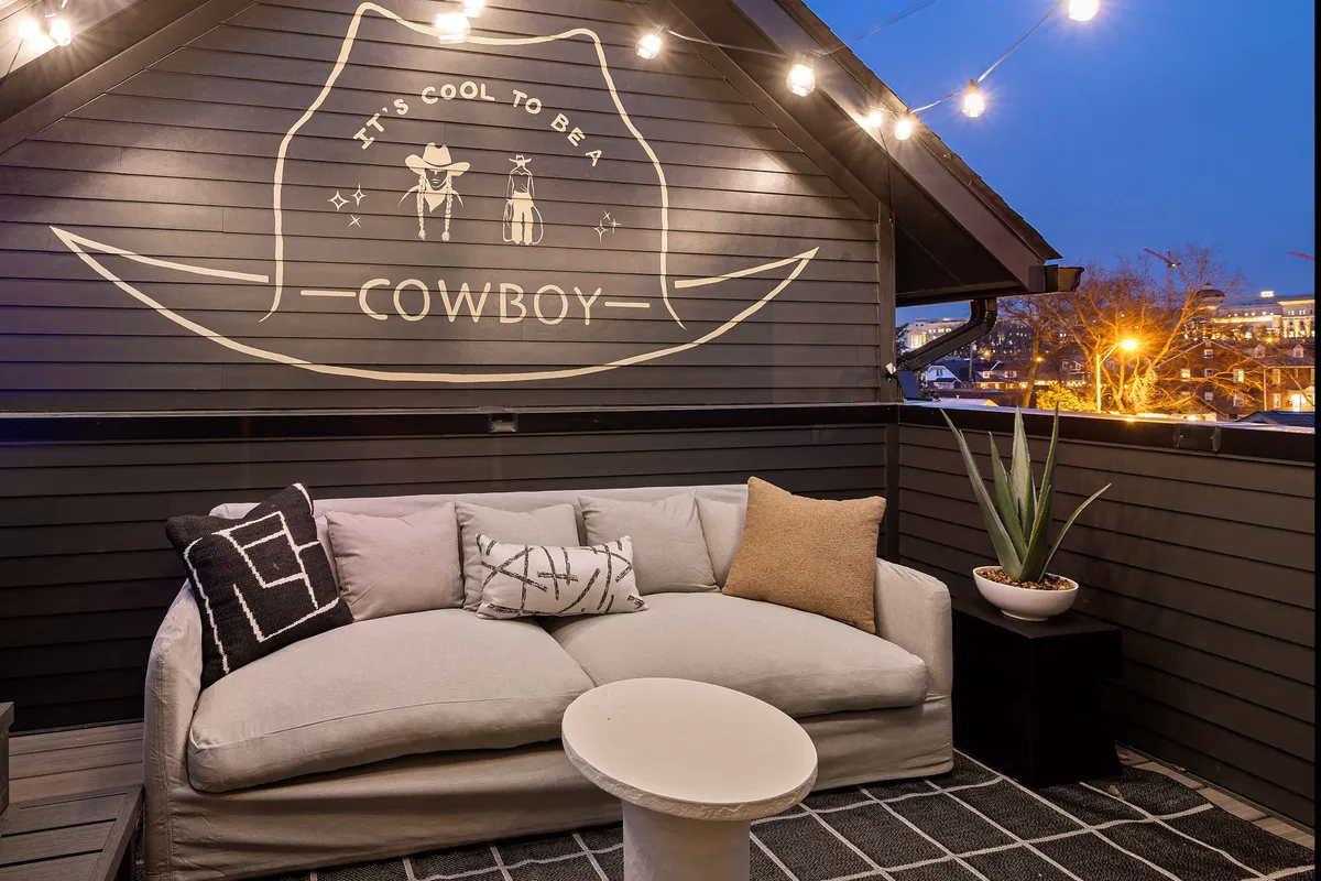 An inviting rooftop outdoor area features a playful cowboy mural, cozy seating under string lights, and a relaxed vibe perfect for socializing or unwinding beneath the stars. The scene suggests an intimate, designer-furnished space ideal for both quiet mornings and lively gatherings.