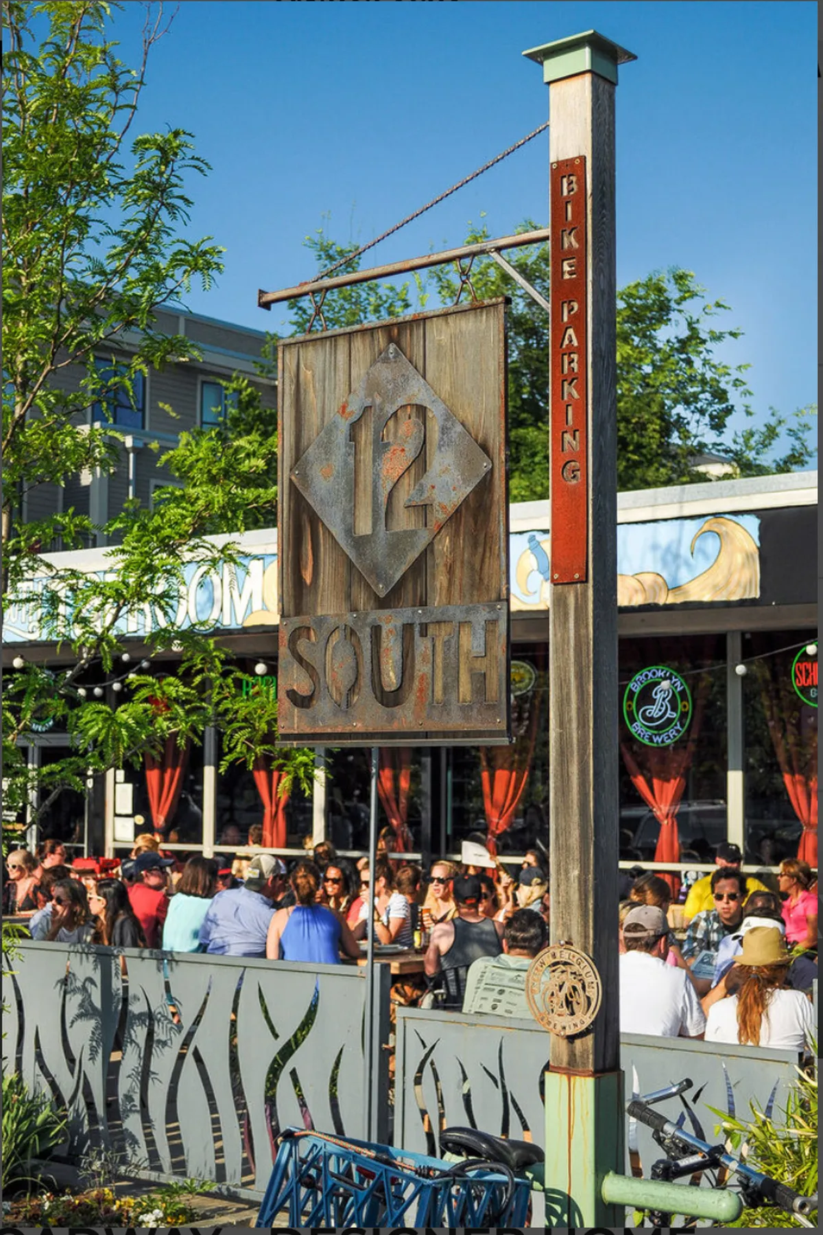 A vibrant street scene in the 12 South neighborhood, with crowds of people walking, dining, and socializing near a large '12 South' sign and bike parking. The energy of the area is palpable, with local eateries and community activity in full swing.