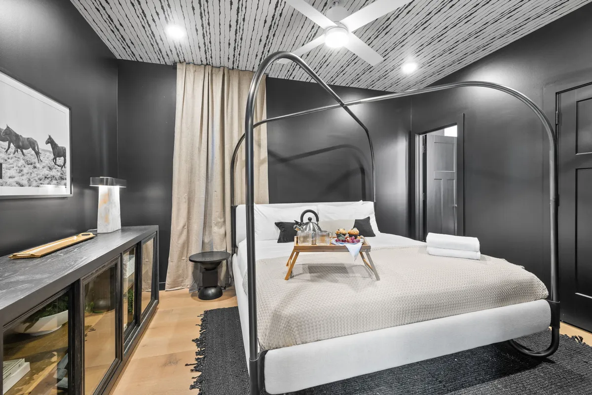 A sunlit bedroom featuring a canopy bed dressed in crisp white linens, a breakfast tray placed invitingly on the bed, and vibrant artwork on the wall. Soft, ambient light streams through the window, creating a cozy and intimate atmosphere perfect for relaxation or a romantic getaway.